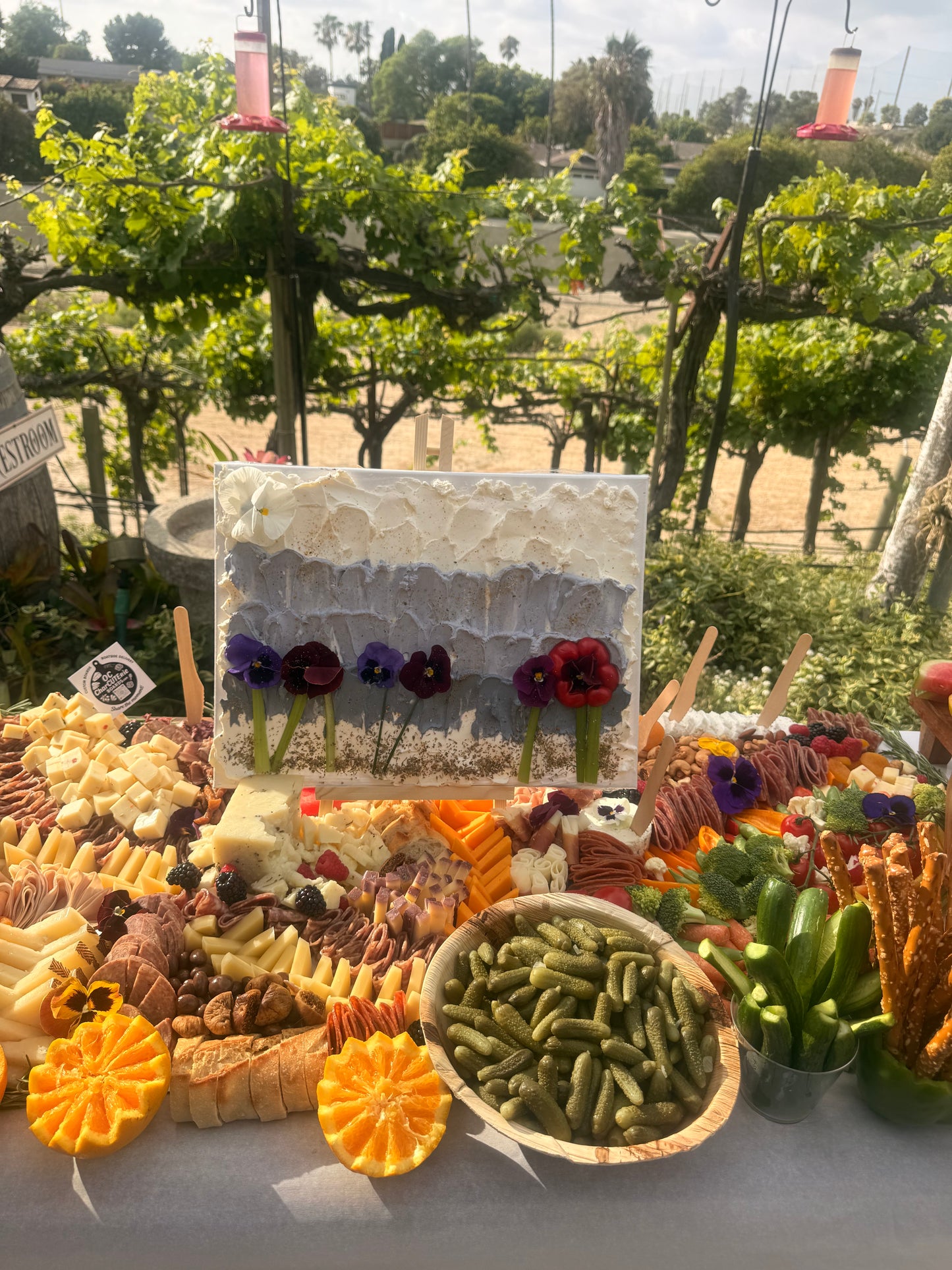 Edible floral cheese canvas on easel beside grazing table spread, OC Charcuterie Boards